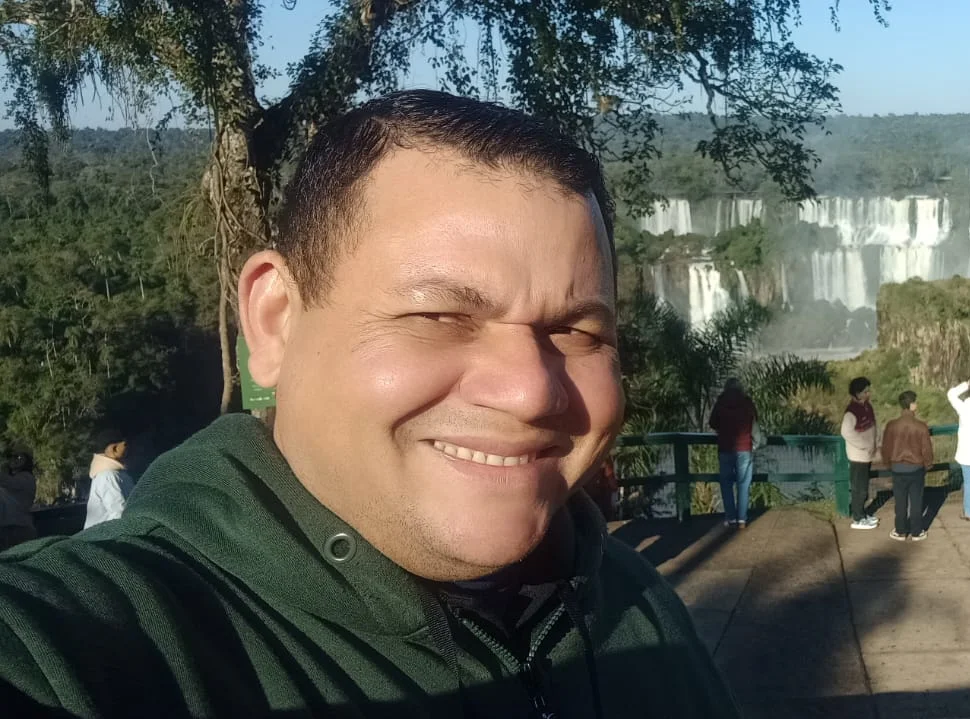 Guia de turismo Reinaldo sorrindo em selfie com as Cataratas do Iguaçu (lado argentino) ao fundo.